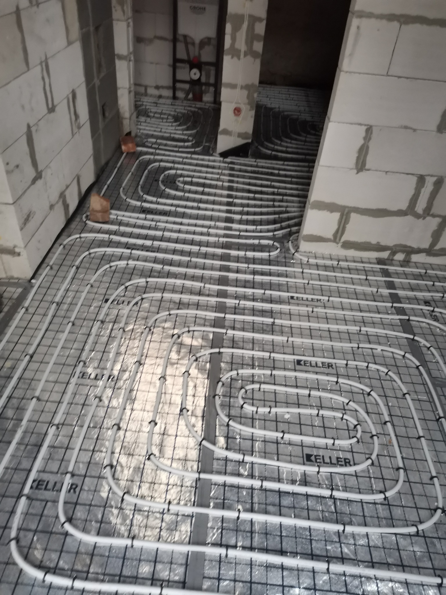 Rozłożone białe rury ogrzewania podłogowego marki Keller na czarnej siatce, widoczne surowe ściany z betonu komórkowego w trakcie prac wykończeniowych.