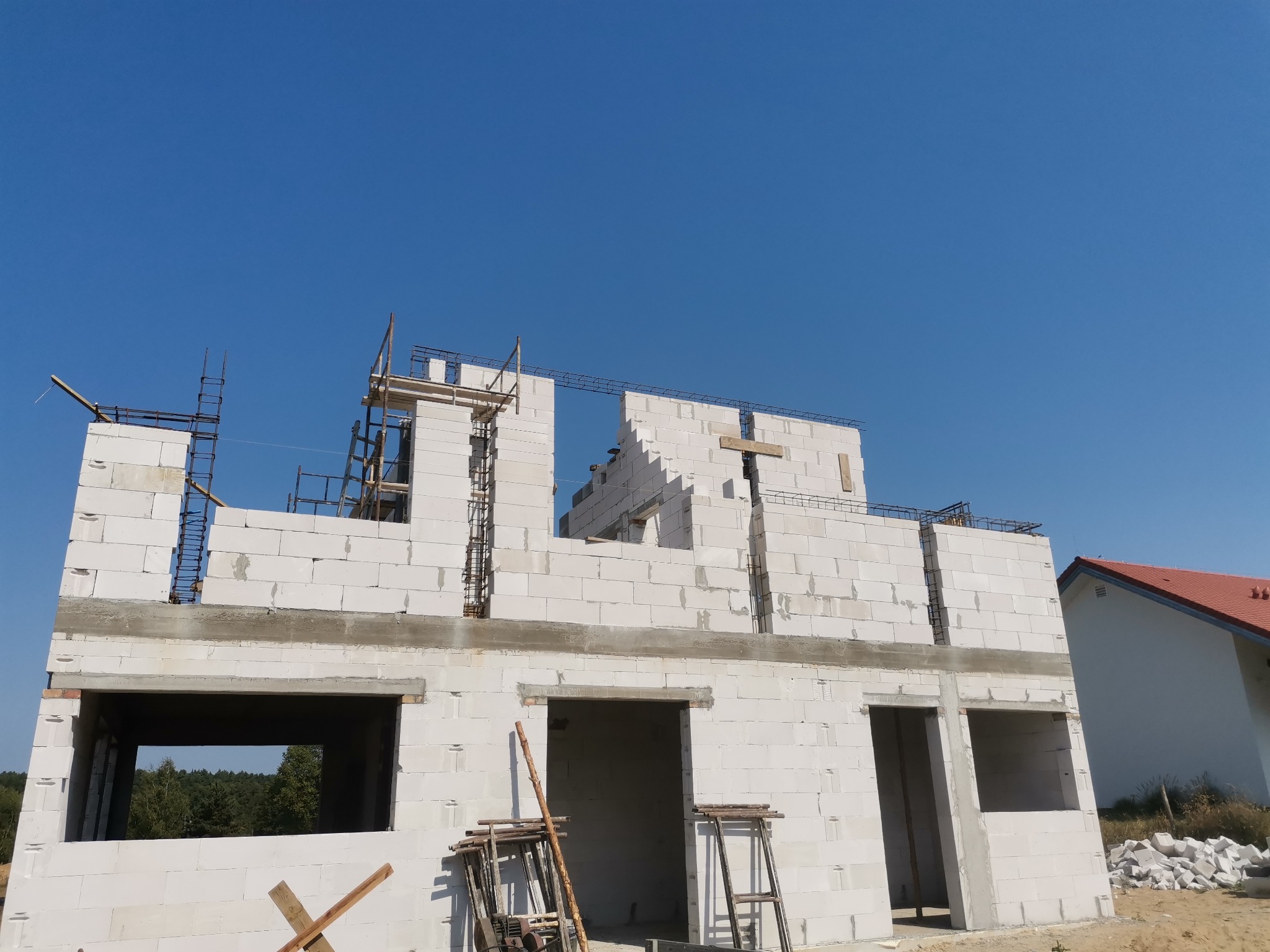 Budowa domu jednorodzinnego z białych bloczków, widoczne zbrojenia i rusztowania, błękitne niebo w tle.