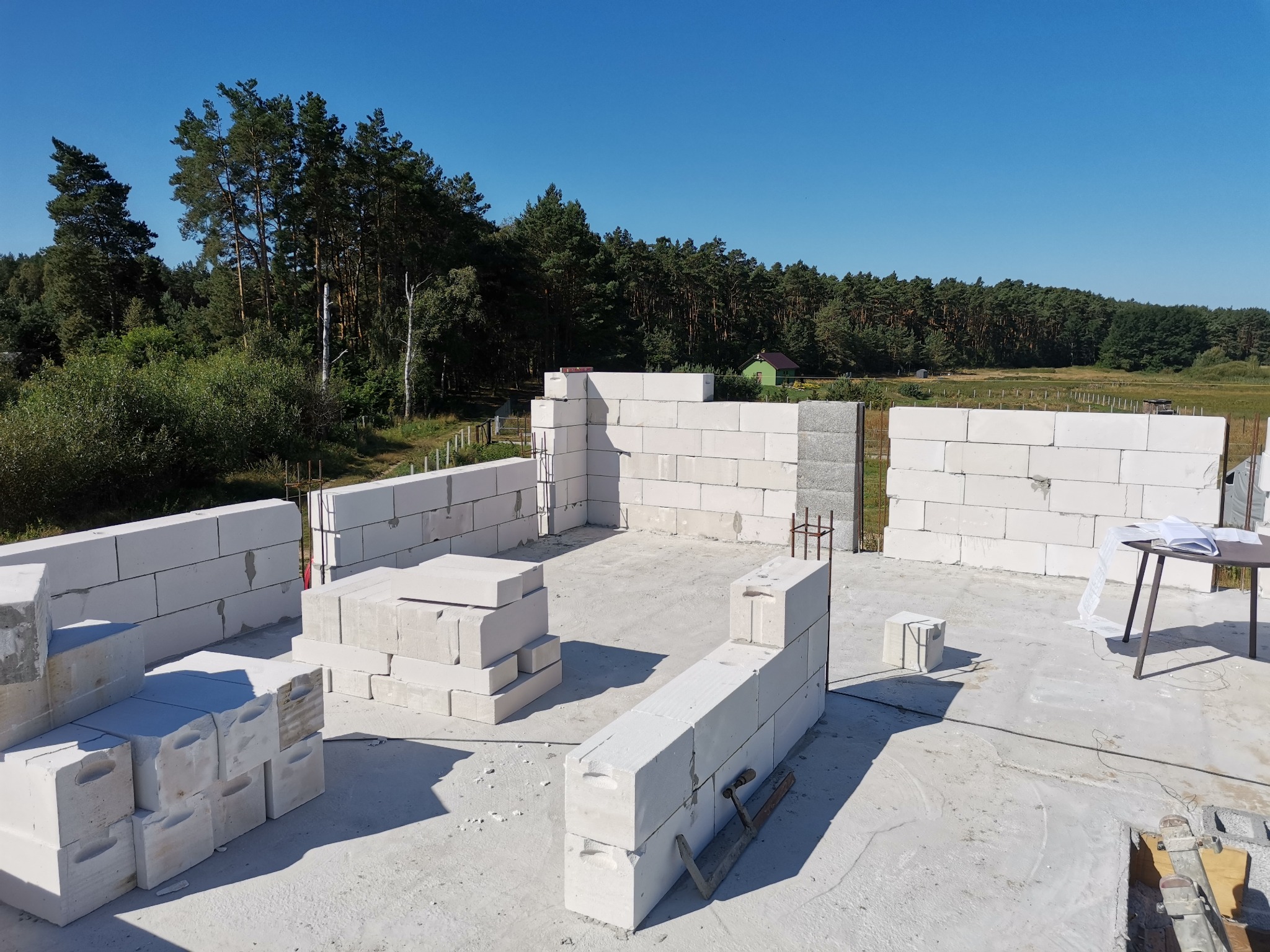 Wznoszone ściany parteru z białych bloczków betonu komórkowego na fundamencie, widoczne narzędzia murarskie i stół z dokumentami, w tle las i błękitne niebo.