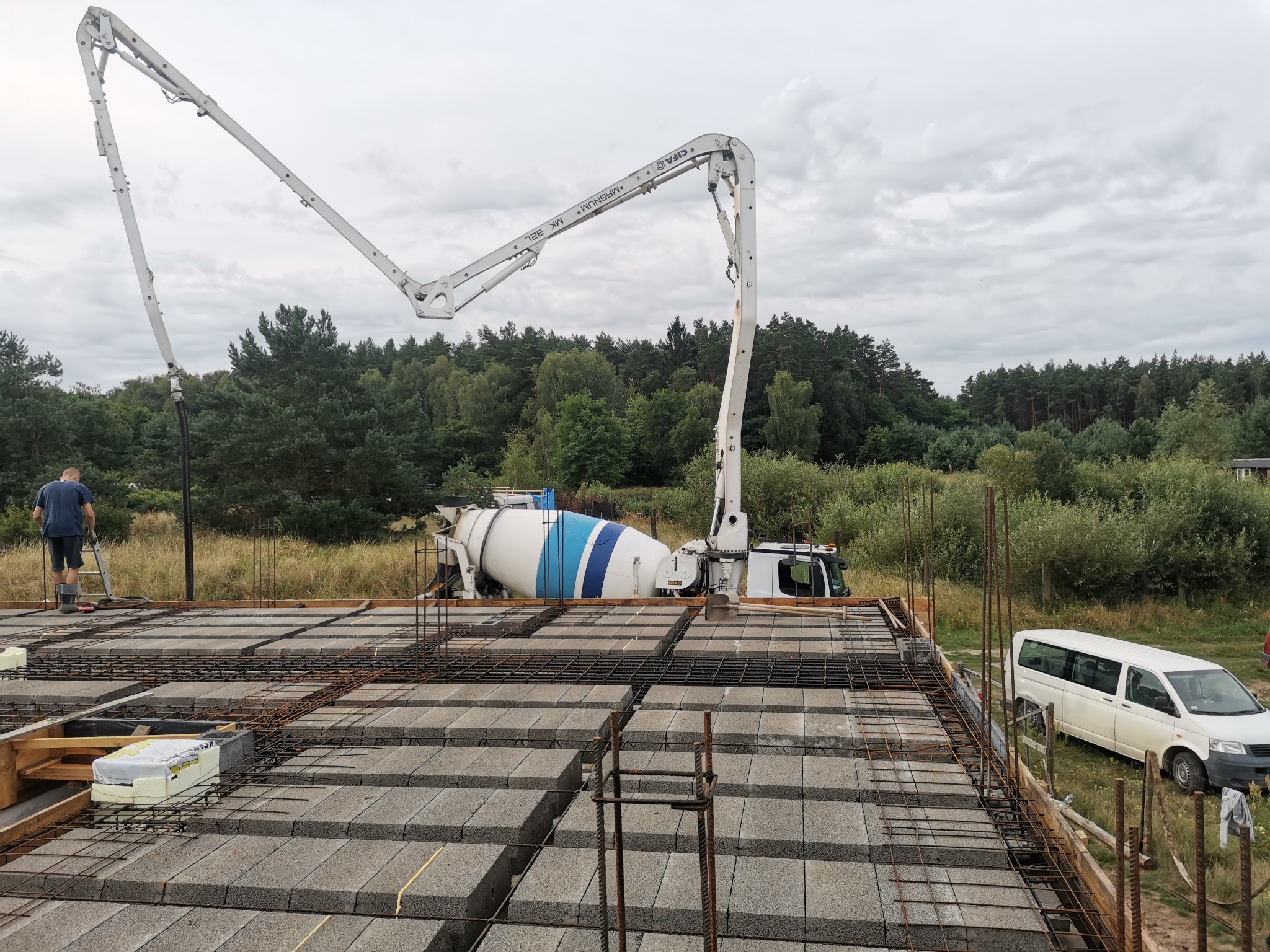 Betoniarka z pompą podaje beton na strop Teriva, widoczne zbrojenie i pustaki szalunkowe, w tle las i pochmurne niebo.