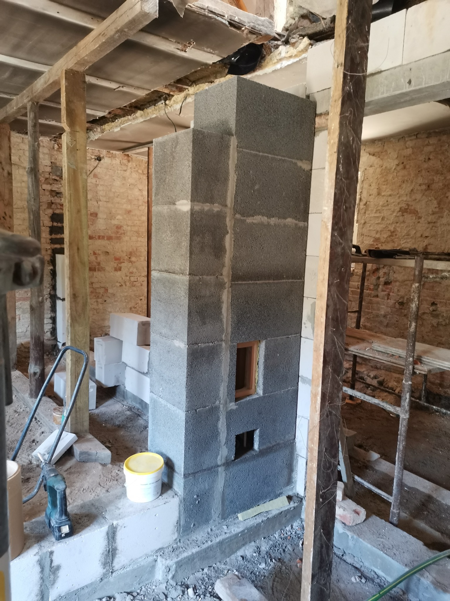 Wznoszenie komina z szarego bloczka betonowego w surowym wnętrzu, widoczne drewniane podpory, rusztowanie, narzędzia i materiały budowlane, etap budowy.