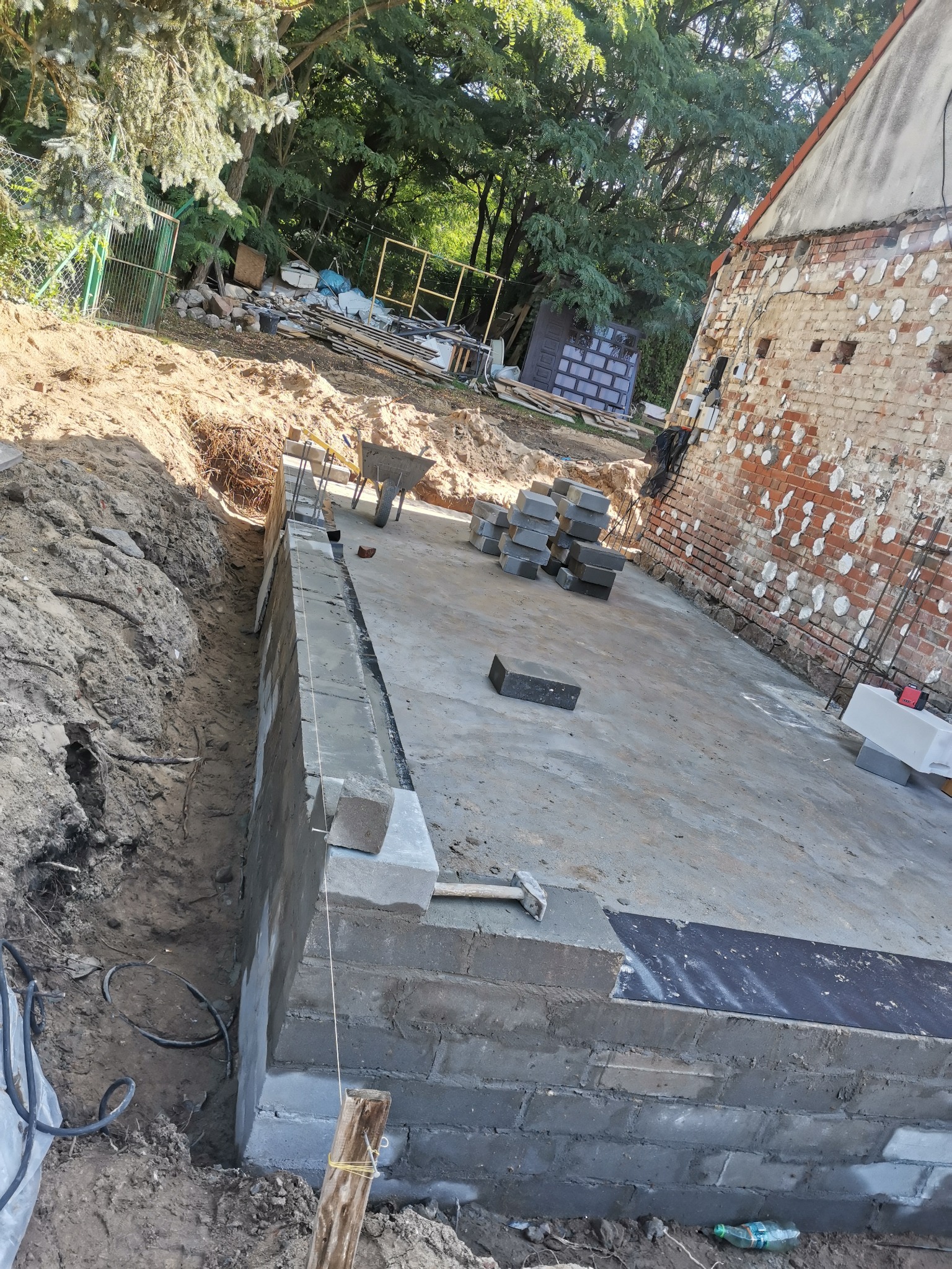 Budowa fundamentów z bloczków betonowych przy ceglanym budynku, widoczny wykop, bloczki ułożone na zaprawie, sznurek murarski i narzędzia.