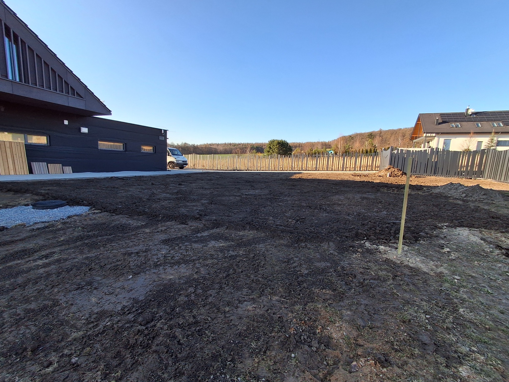 Przygotowany, wyrównany teren pod trawnik lub ogród, widoczny fragment nowoczesnego domu z ciemną elewacją, biały van zaparkowany w tle, słoneczny dzień.