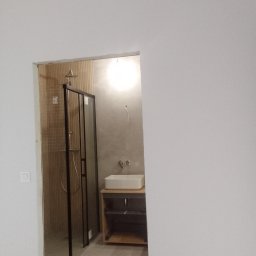 Stanisław Czarnecki - Nowoczesna łazienka w trakcie wykańczania: kabina prysznicowa z czarnymi ramami, umywalka nablatowa na drewnianej szafce, szare ściany i drewnopodobne płytki w strefie prysznica.