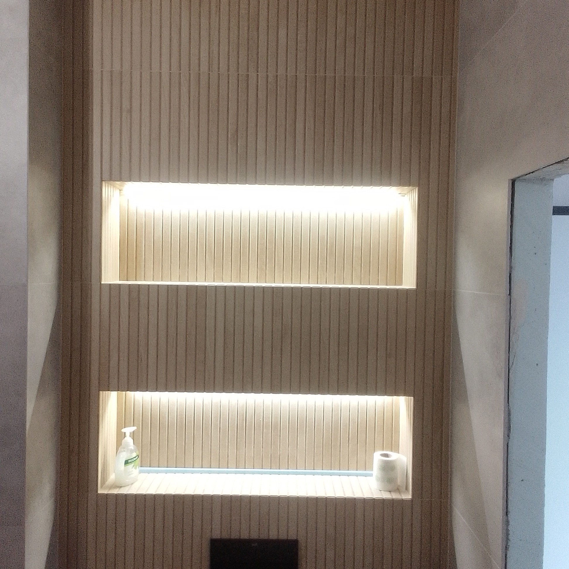 Pionowe panele ścienne z dwoma podświetlonymi wnękami, w jednej mydło w płynie, w drugiej rolka papieru toaletowego, nowoczesna aranżacja łazienki.