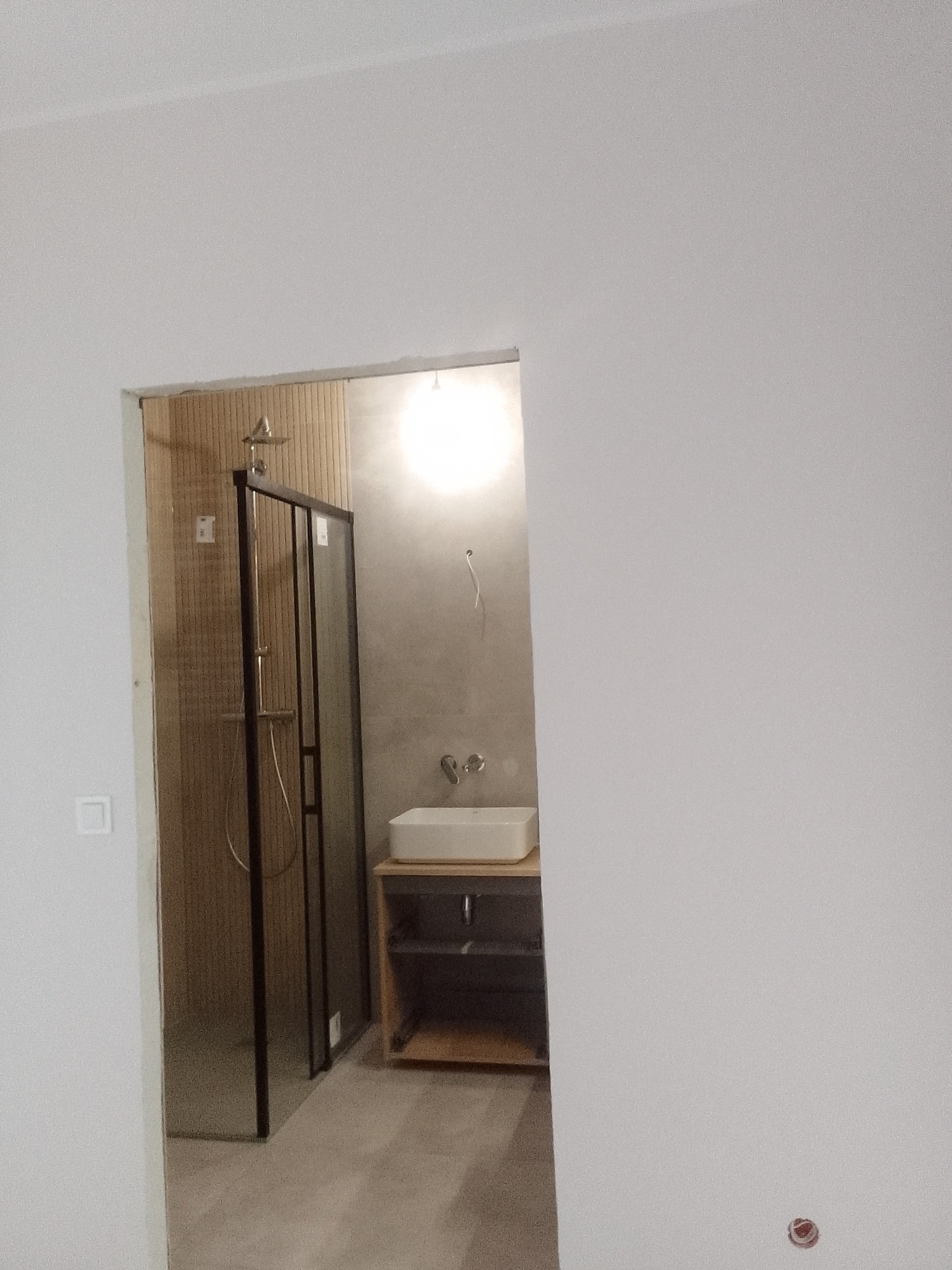 Nowoczesna łazienka w trakcie wykańczania: kabina prysznicowa z czarnymi ramami, umywalka nablatowa na drewnianej szafce, szare ściany i drewnopodobne płytki w strefie prysznica.