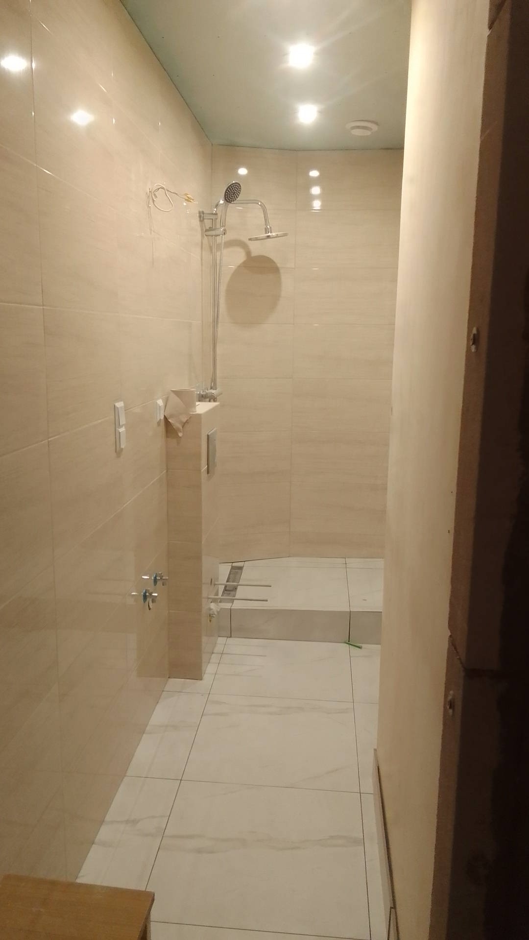 Jasna łazienka w trakcie prac wykończeniowych, widoczne płytki imitujące marmur na ścianach i podłodze, zamontowana bateria prysznicowa, wystające przewody elektryczne oraz niedokończona zabudowa...