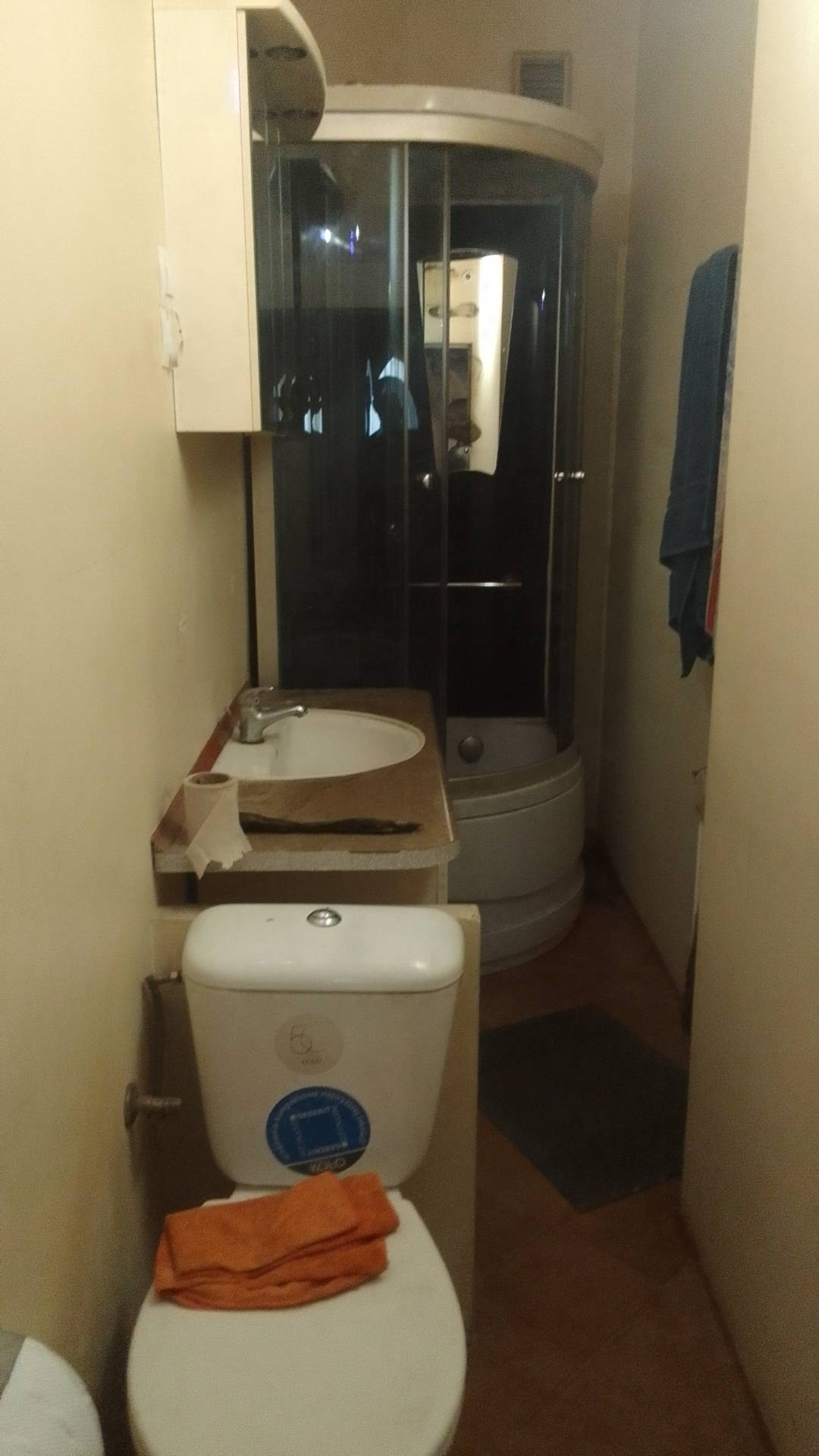 Niewielka łazienka z kabiną prysznicową, umywalką w trakcie montażu, toaletą z pomarańczowym ręcznikiem i szarą wycieraczką na podłodze.