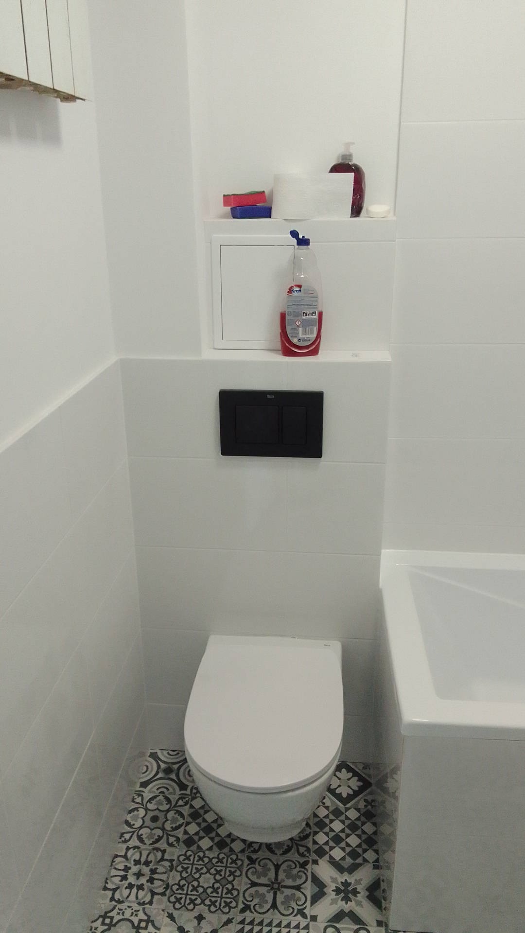 Biała toaleta wisząca na tle wzorzystych, czarno-białych płytek podłogowych, z widoczną częścią wanny i przyciskiem spłukującym w kolorze czarnym, a także środkami czystości na zabudowie geberit