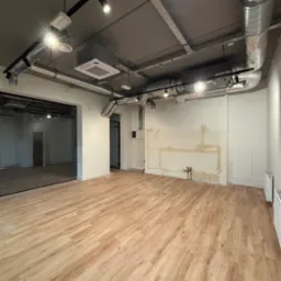 Wnętrze lokalu w trakcie prac instalacyjnych: widoczne kanały wentylacyjne pod sufitem, instalacja elektryczna, fragmenty instalacji wodnej na ścianie, nowa podłoga imitująca drewno.