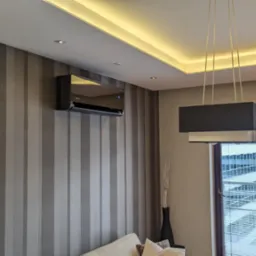 Elegancki, czarny klimatyzator ścienny zamontowany w salonie we Wrocławiu z tapetą w pionowe pasy, oświetleniem LED pod sufitem i nowoczesną lampą wiszącą.