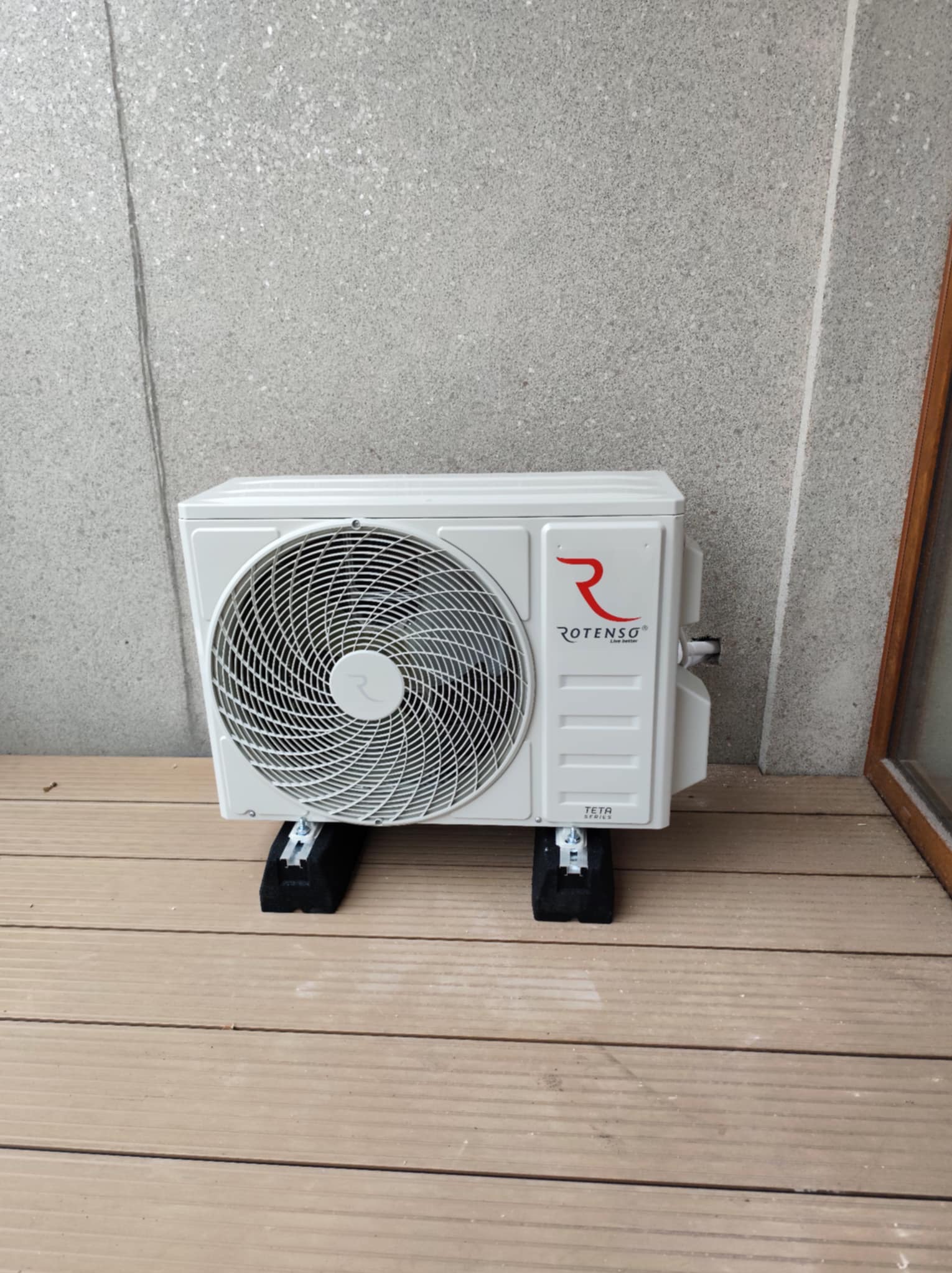 Biała jednostka zewnętrzna klimatyzatora Rotenso Teta Series, umieszczona na czarnych wspornikach na drewnianym tarasie, na tle szarej ściany.