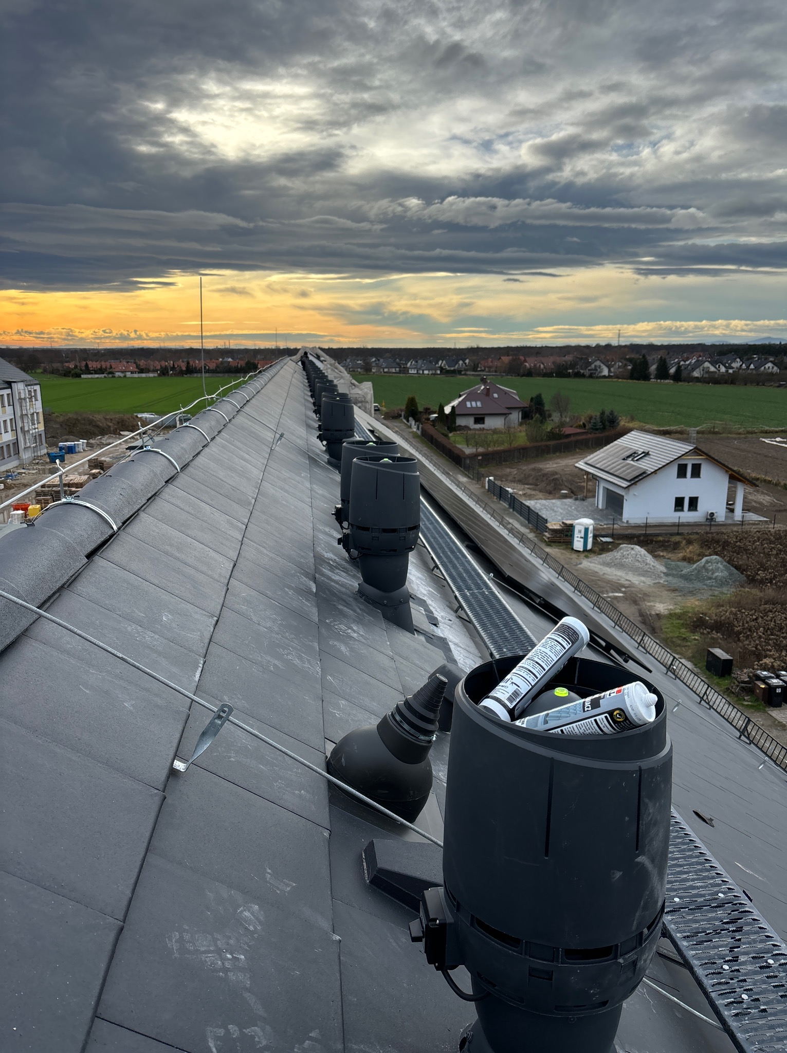 Szereg czarnych kominków wentylacyjnych na szarym dachu z dachówki, widok z góry na okoliczne domy i zachodzące słońce, w jednym z kominków tuby z uszczelniaczem.