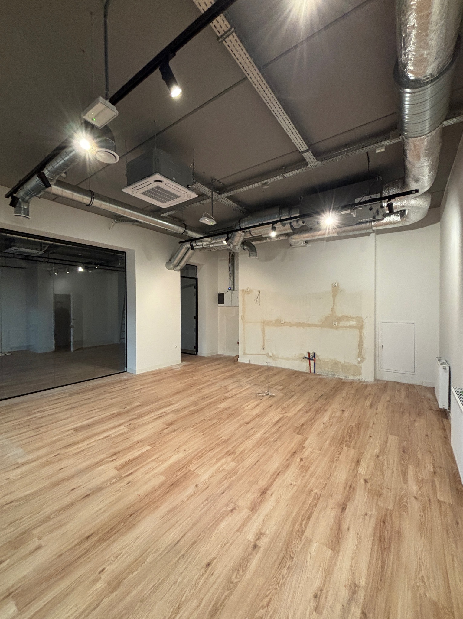 Wnętrze lokalu w trakcie prac instalacyjnych: widoczne kanały wentylacyjne pod sufitem, instalacja elektryczna, fragmenty instalacji wodnej na ścianie, nowa podłoga imitująca drewno.
