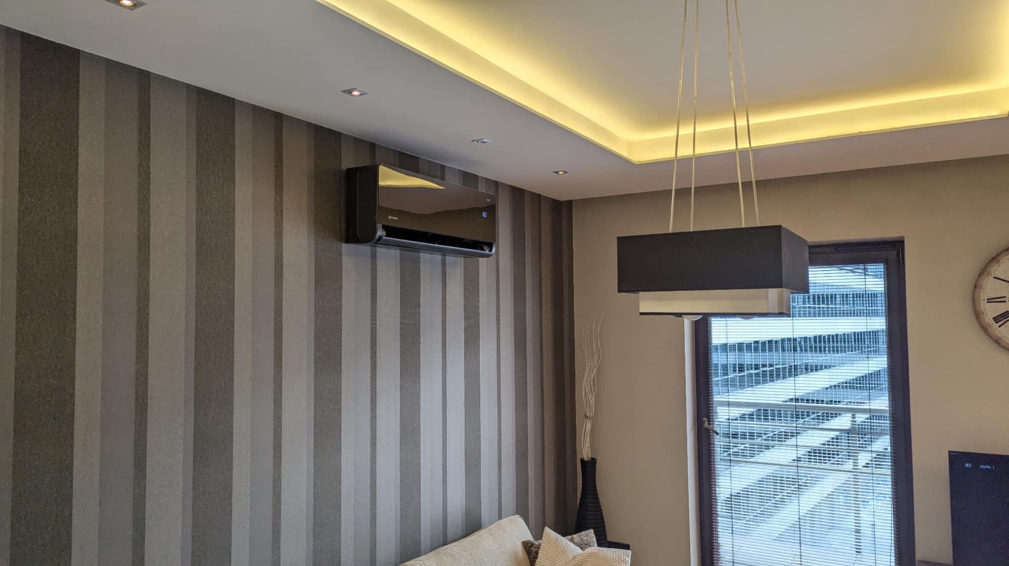 Elegancki, czarny klimatyzator ścienny zamontowany w salonie we Wrocławiu z tapetą w pionowe pasy, oświetleniem LED pod sufitem i nowoczesną lampą wiszącą.