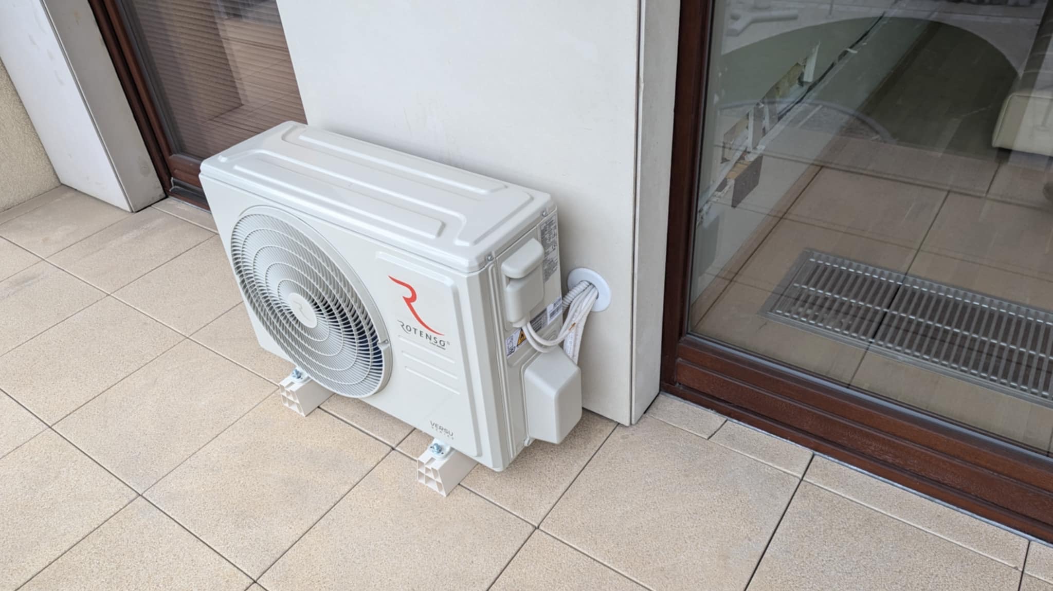 Biała jednostka zewnętrzna klimatyzacji marki Rotenso, zamontowana na balkonie z beżowymi płytkami, widoczne przewody wchodzące do ściany przez otwór z uszczelką.