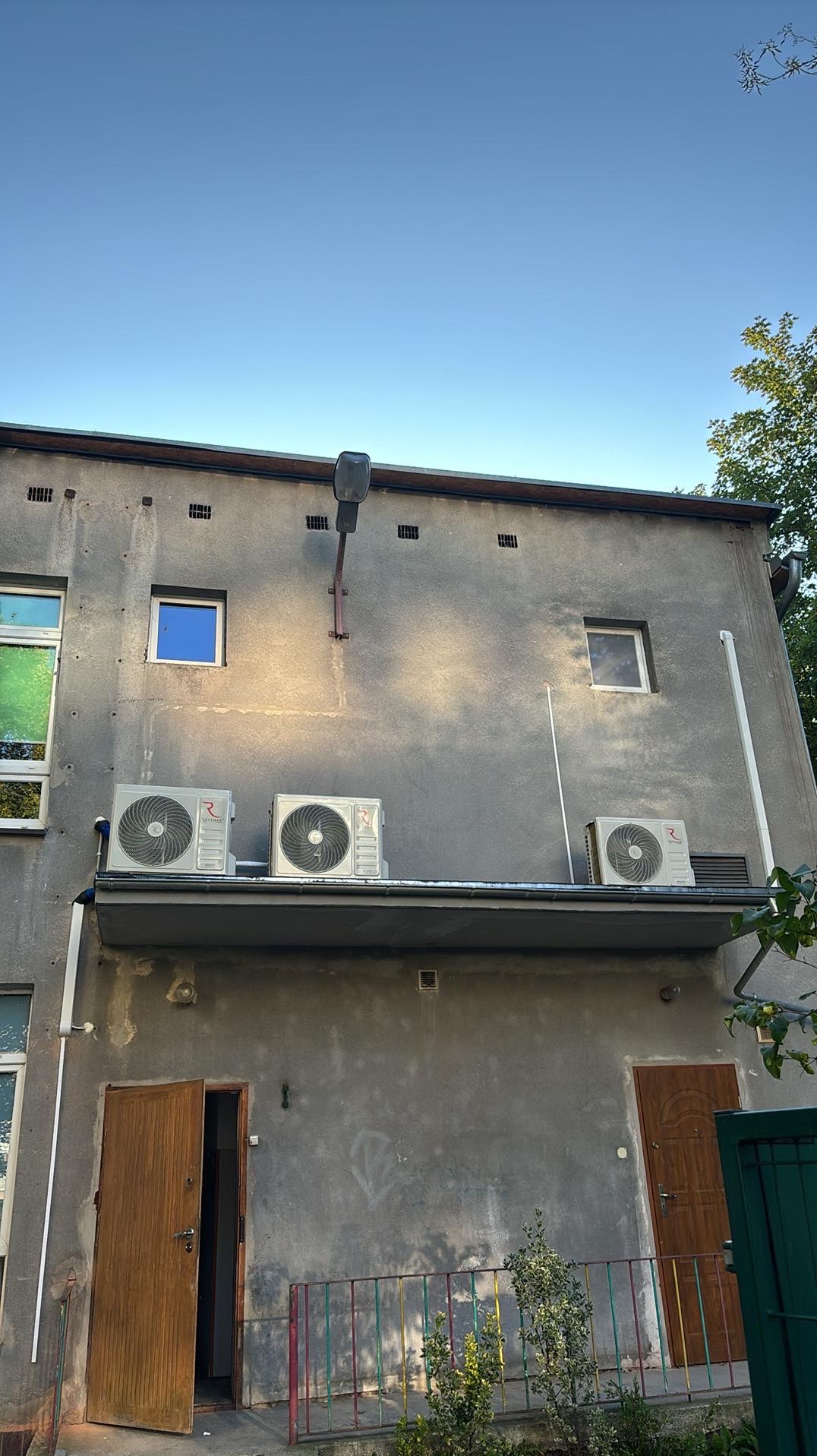 Trzy jednostki klimatyzacyjne zamontowane na zadaszeniu szarego budynku z widocznymi oknami i otwartymi drzwiami wejściowymi, oświetlone popołudniowym słońcem.