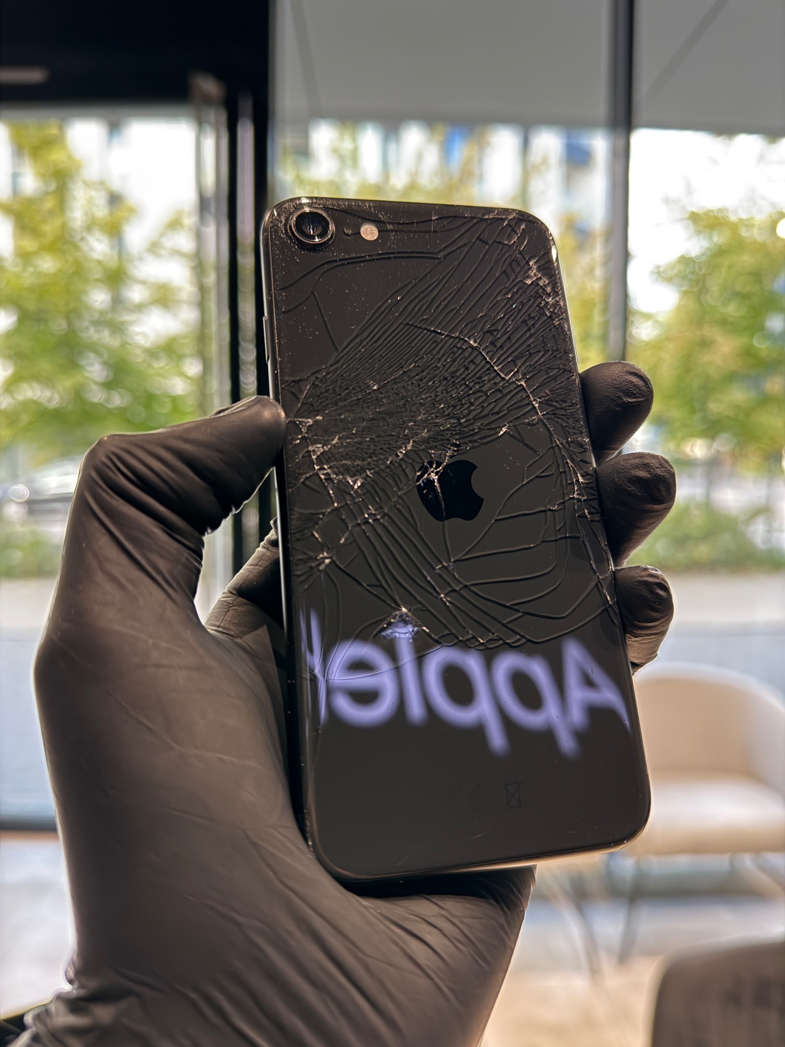 Czarny iPhone z pękniętą szybą z tyłu, trzymany w dłoni w czarnej rękawiczce, widoczne logo Apple i odbicie napisu 'Apple' na powierzchni.