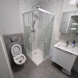 Dmytro Tkachuk - Wykończona łazienka z kabiną prysznicową, umywalką z szafką i toaletą. Na ścianach szare płytki z geometrycznym wzorem. Podłoga wyłożona szarymi płytkami.