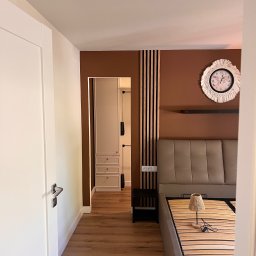 Dmytro Tkachuk - Nowoczesna sypialnia z brązową ścianą, drewnianymi lamelami i dekoracyjnym zegarem. Widoczny fragment łóżka i drzwi do garderoby z białymi meblami.