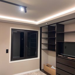 Dmytro Tkachuk - Nowoczesna garderoba z ciemnymi meblami, oświetleniem LED i oknem. Widoczny fragment dywanu i walizki. Minimalistyczny design wnętrza.