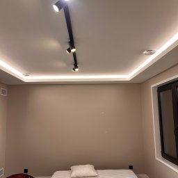 Dmytro Tkachuk - Beżowa ściana w pokoju z listwami LED pod sufitem i reflektorami na szynie. Na podłodze materac i walizki. Widoczne okno z ciemną ramą.