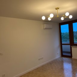 Dmytro Tkachuk - Jasne wnętrze po odświeżeniu: parkiet, białe ściany z listwami przypodłogowymi, nowoczesny żyrandol i klimatyzator. Widok na balkon przez drewniane drzwi.