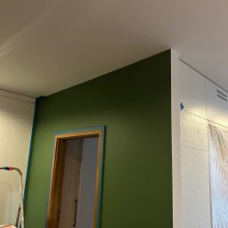 Dmytro Tkachuk - Ściana pomalowana na zielono z zabezpieczonymi taśmą malarską drzwiami i folią meblami. Widoczna drabina malarska. Świeżo pomalowane wnętrze w trakcie prac wykończeniowych.
