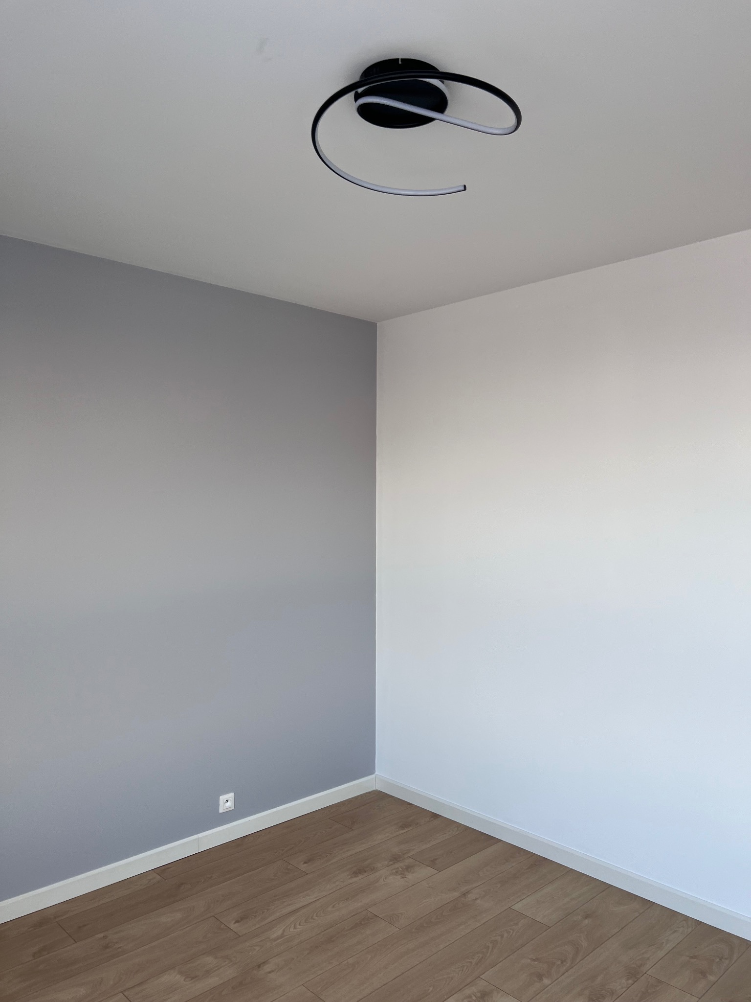 Pomalowany pokój z drewnianą podłogą, jedna ściana szara, druga biała. Na suficie nowoczesna lampa. Widoczny fragment listwy przypodłogowej i gniazdko elektryczne.