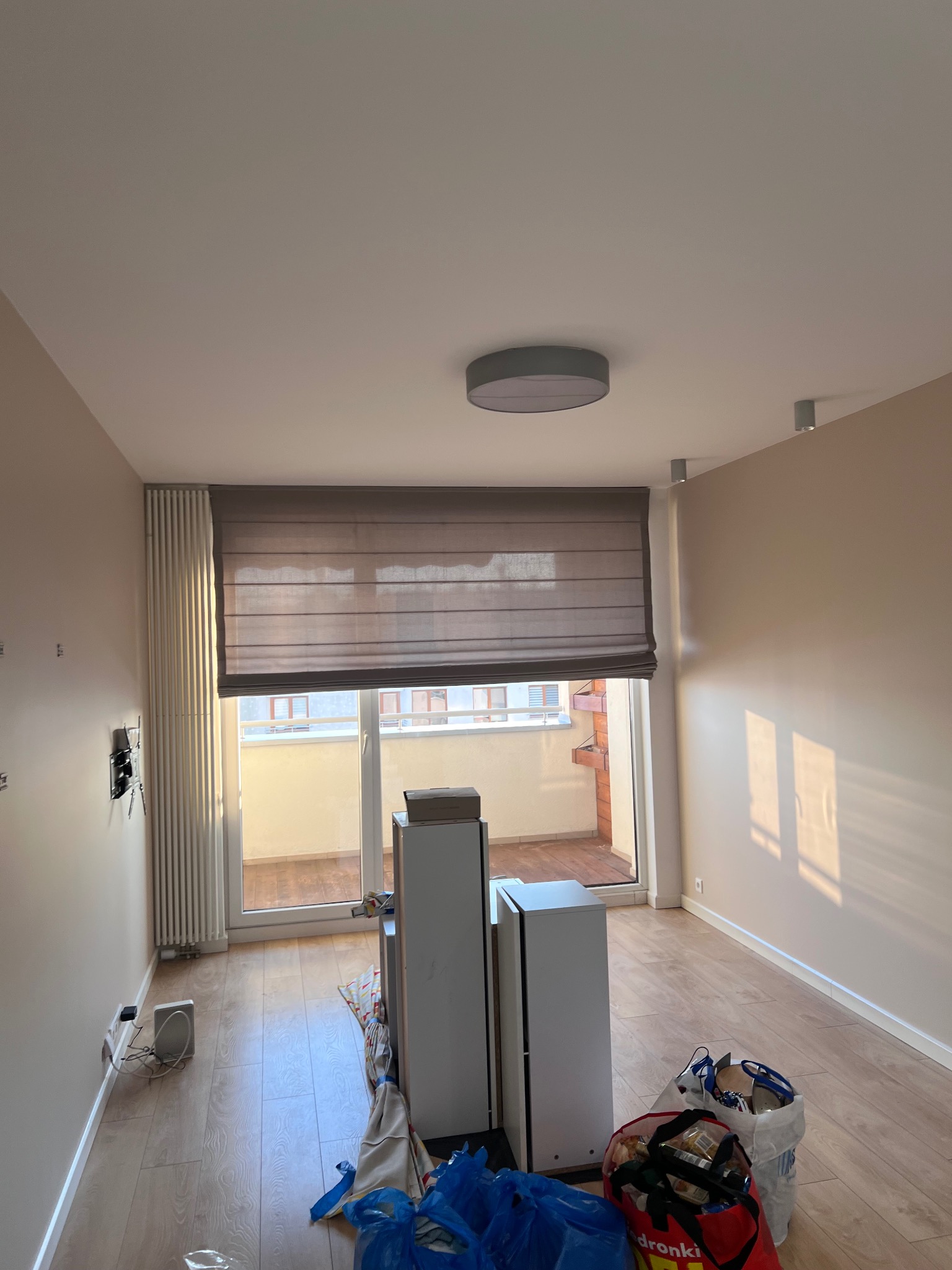 Jasne wnętrze po malowaniu ścian na beżowo, z widocznymi śladami prac remontowych: worki, kartony, odsłonięte gniazdka. Parkiet na podłodze, okno balkonowe z roletą rzymską.