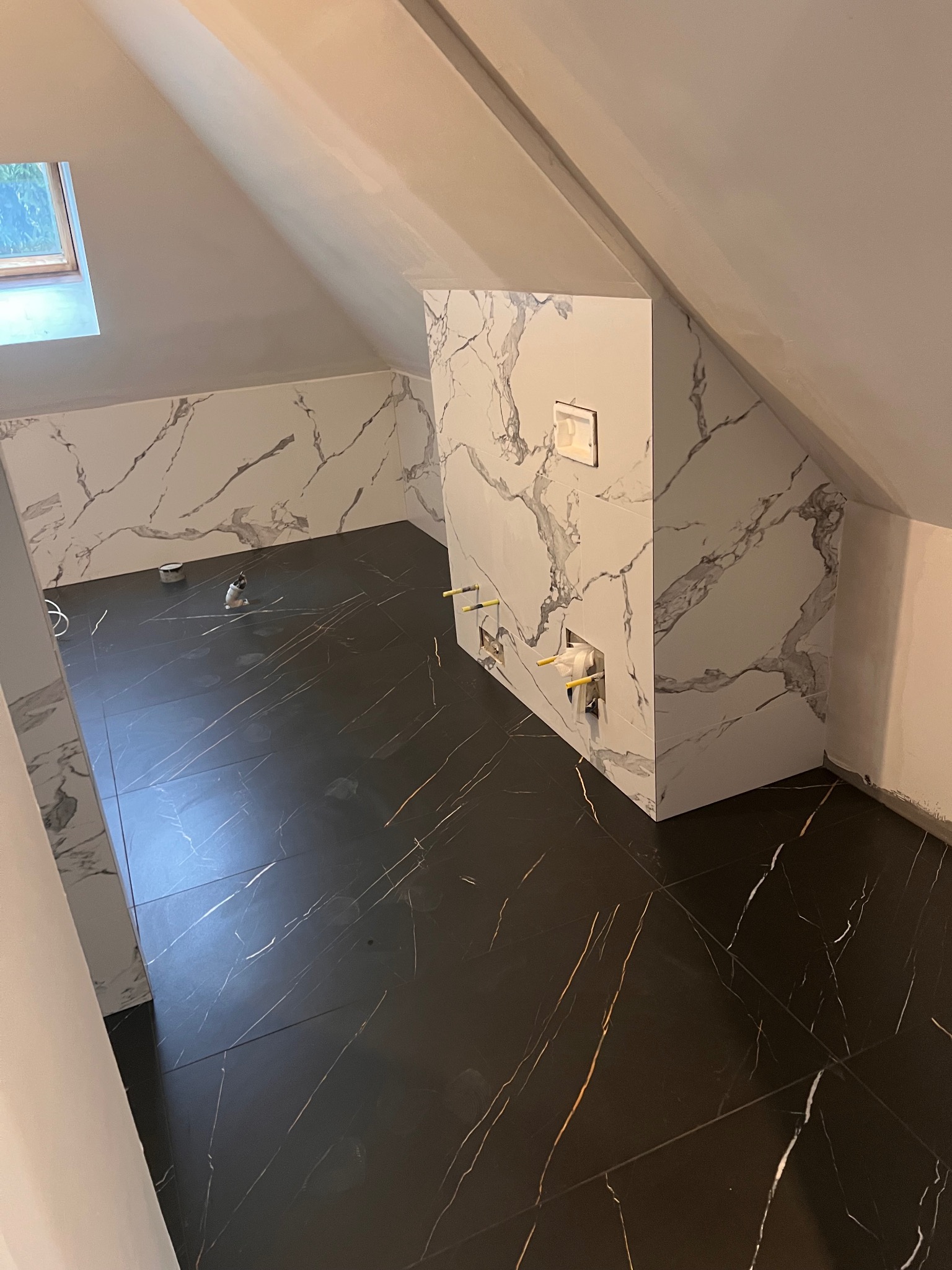 Wykończona łazienka na poddaszu z czarną podłogą w złote żyły i ścianami w białe marmurowe płyty. Widoczne przyłącza wodne i okno dachowe.