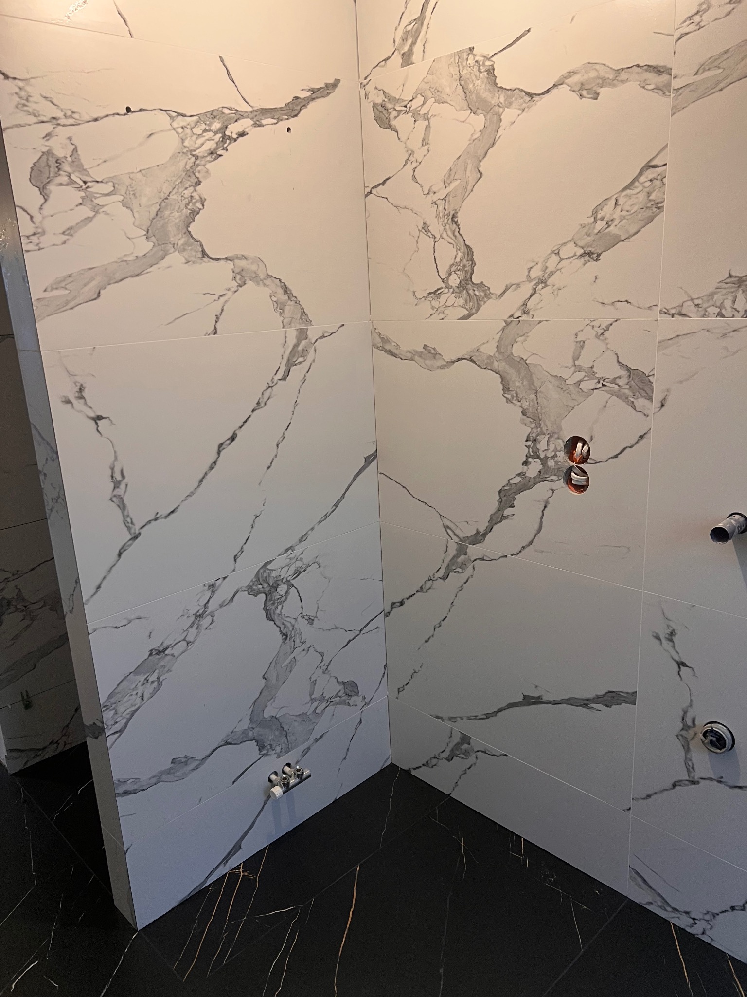 Wykończona łazienka z czarno-białymi płytkami imitującymi marmur na ścianach i podłodze. Widoczne przyłącza wodne i kanalizacyjne.