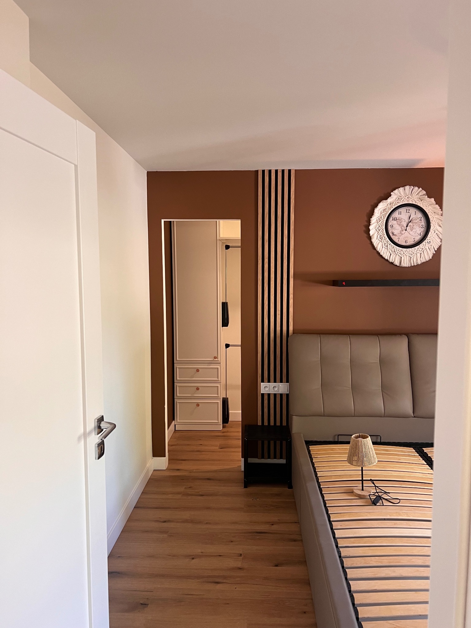 Nowoczesna sypialnia z brązową ścianą, drewnianymi lamelami i dekoracyjnym zegarem. Widoczny fragment łóżka i drzwi do garderoby z białymi meblami.
