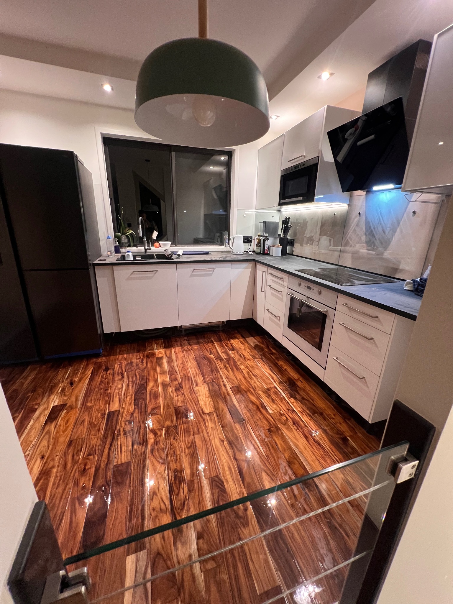 Błyszcząca, drewniana podłoga w kuchni z białymi szafkami i czarną lodówką. Widok z góry schodów z szklaną balustradą. Nowoczesne oświetlenie.