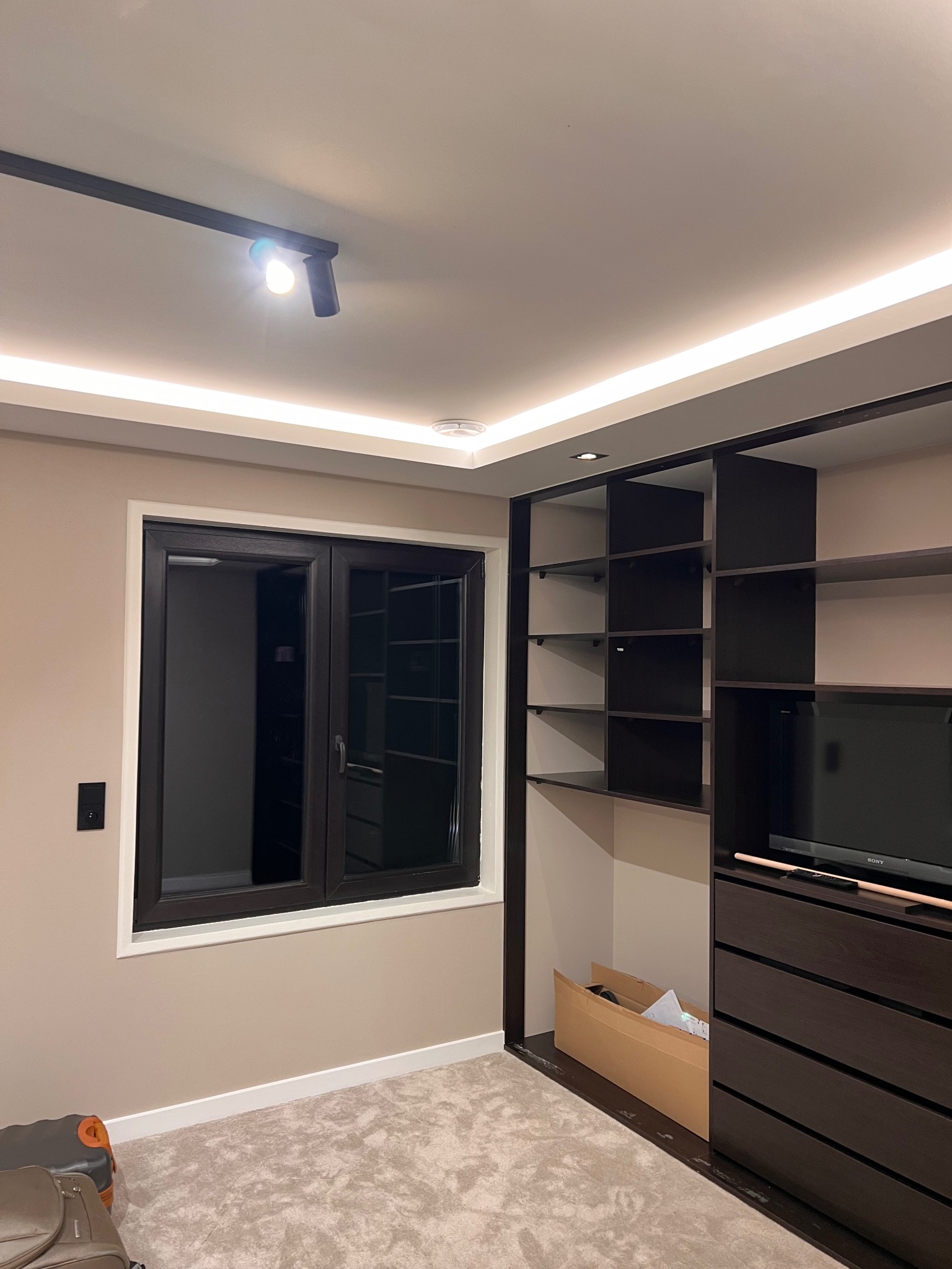 Nowoczesna garderoba z ciemnymi meblami, oświetleniem LED i oknem. Widoczny fragment dywanu i walizki. Minimalistyczny design wnętrza.