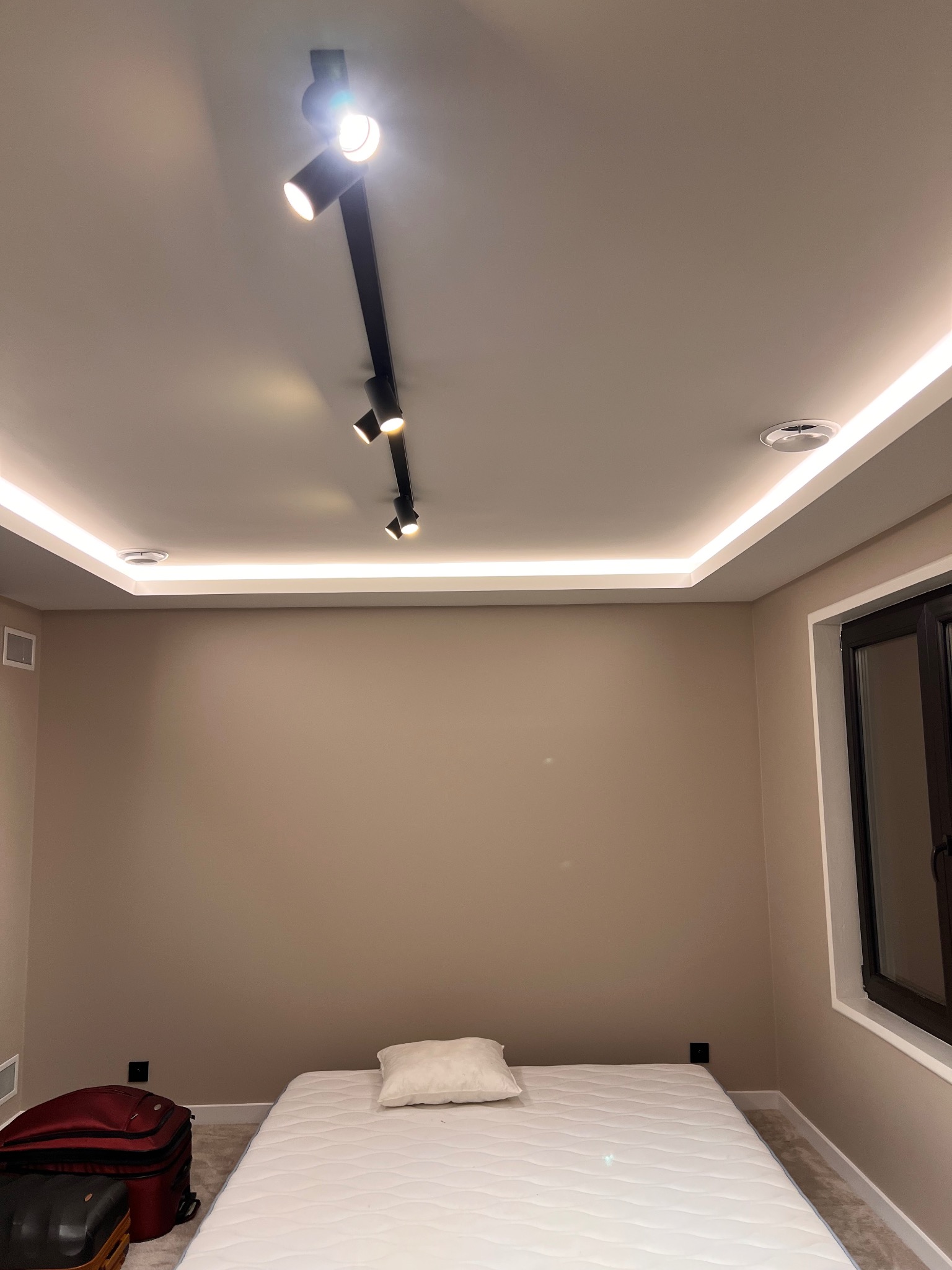 Beżowa ściana w pokoju z listwami LED pod sufitem i reflektorami na szynie. Na podłodze materac i walizki. Widoczne okno z ciemną ramą.
