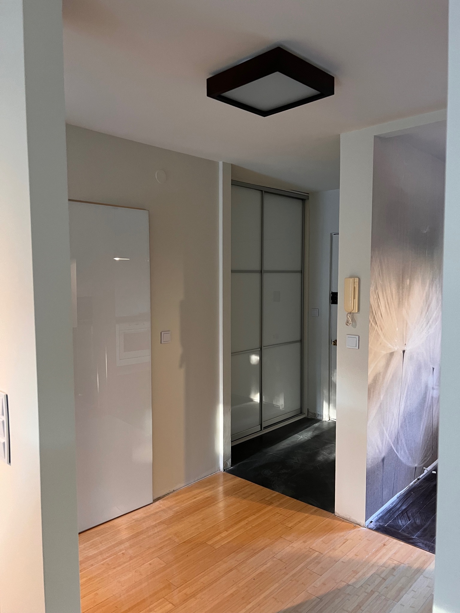 Jasne wnętrze mieszkania po remoncie: parkiet, białe ściany, przesuwne drzwi do garderoby, nowoczesna lampa sufitowa. Częściowo zakryte folią zabezpieczającą.