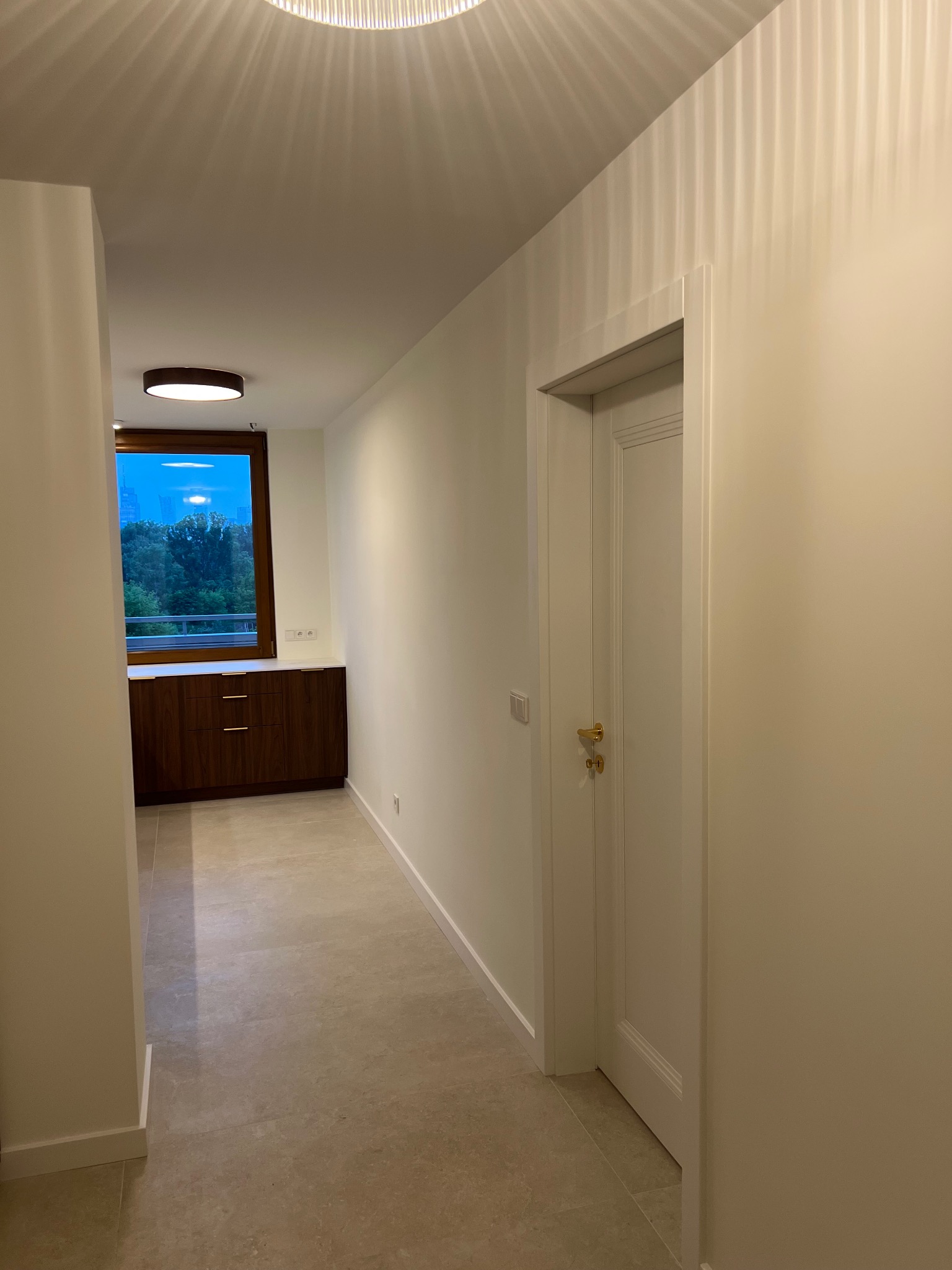Jasny korytarz z beżową podłogą, białymi ścianami i drzwiami. W tle okno z widokiem na zieleń i zabudowa meblowa. Nowoczesne oświetlenie sufitowe.