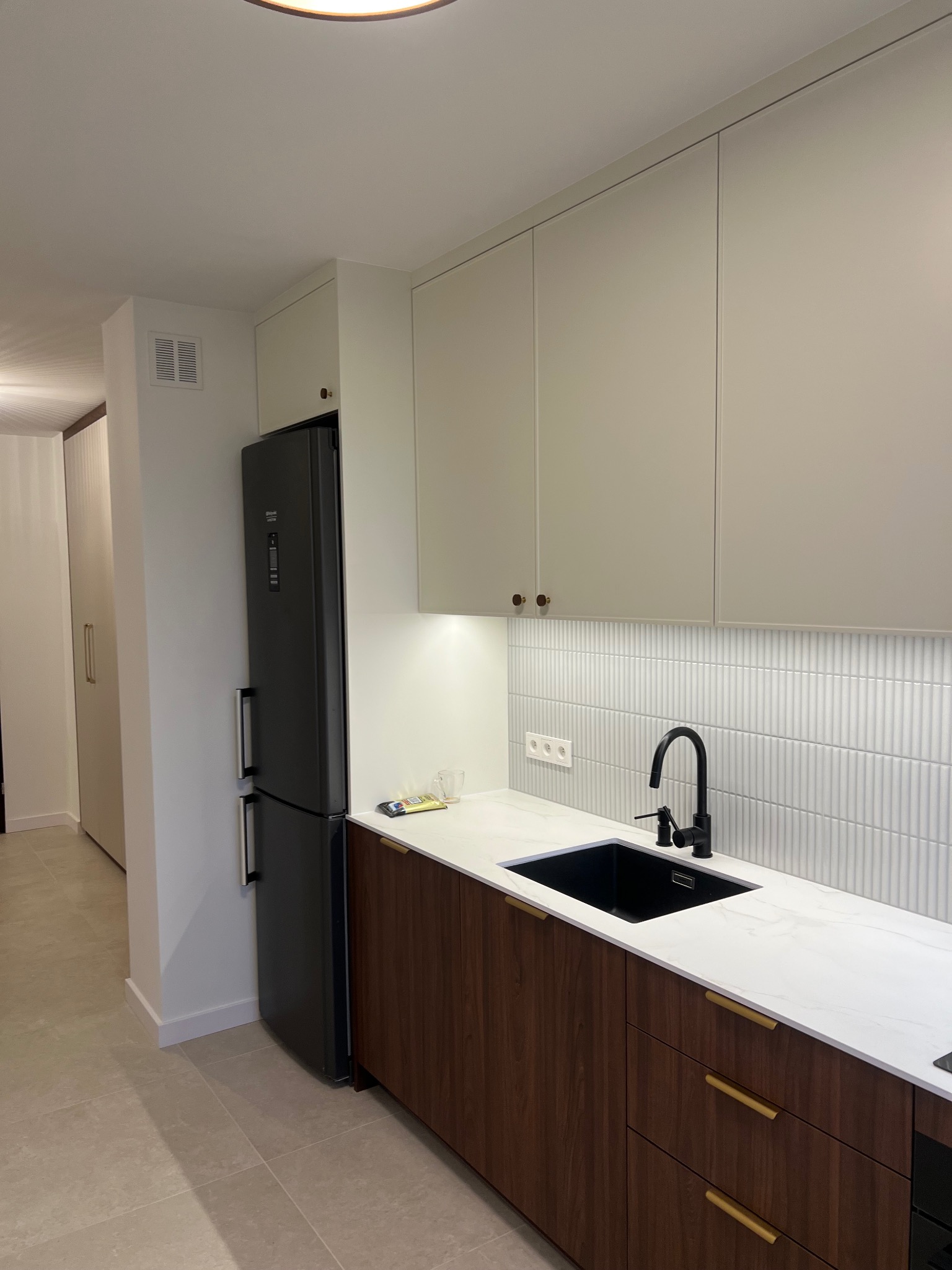 Nowoczesna kuchnia z białymi szafkami, drewnianymi frontami dolnymi i czarną armaturą. Uwagę przyciąga pionowa, biała glazura na ścianie nad blatem.