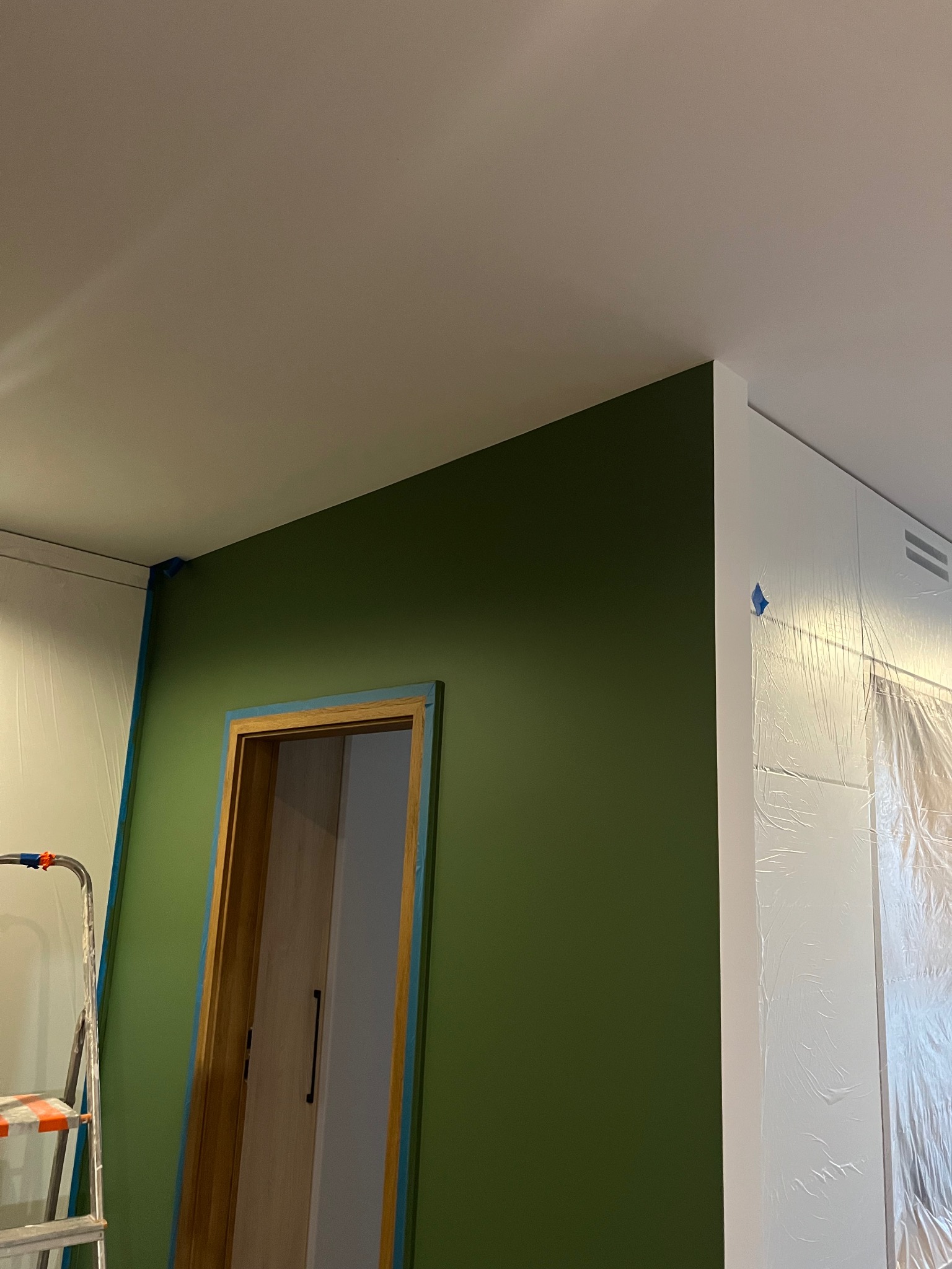 Ściana pomalowana na zielono z zabezpieczonymi taśmą malarską drzwiami i folią meblami. Widoczna drabina malarska. Świeżo pomalowane wnętrze w trakcie prac wykończeniowych.