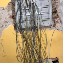 Rozdzielnia elektryczna w trakcie montażu w ścianie z żółtą farbą i odsłoniętą cegłą, z widocznymi przewodami w kolorach niebieskim, zielonym i brązowym, oraz białymi kablami w listwach.