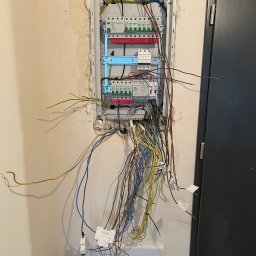 Yurii Tukalo Yurii_bud - Otwarta skrzynka elektryczna w trakcie montażu, widoczne wystające przewody o różnych kolorach, ściana z widocznymi śladami po kuciu, obok ciemne drzwi.