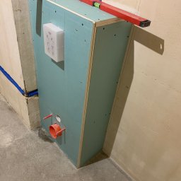 Yurii Tukalo Yurii_bud - Stelaż podtynkowy zabudowany płytą gipsowo-kartonową, poziomowany czerwoną poziomicą, widoczne przyłącza kanalizacyjne i wodne, biały panel sterujący z instrukcjami