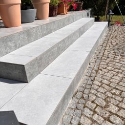 Yurii Tukalo Yurii_bud - Schody zewnętrzne wyłożone jasnoszarą płytką, z donicami z roślinami na górnym stopniu. Obok bruk z kamienia polnego. Słoneczny dzień, widok z góry.