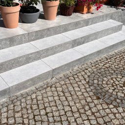Yurii Tukalo Yurii_bud - Schody zewnętrzne wyłożone szarymi płytkami, prowadzące do ogrodu z kostką brukową. Na górze schodów donice z kwiatami.