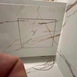Yurii Tukalo Yurii_bud - Białe płytki imitujące marmur z ukrytą klapą rewizyjną w łazience. Widoczny fragment instalacji. Minimalistyczny design z delikatnymi żyłkami.