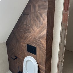 Yurii Tukalo Yurii_bud - Wykończona toaleta w poddaszu z brązową, wzorzystą okładziną ścienną i białym sanitariatem. Widoczny fragment surowej ściany, podkreślający etap prac.