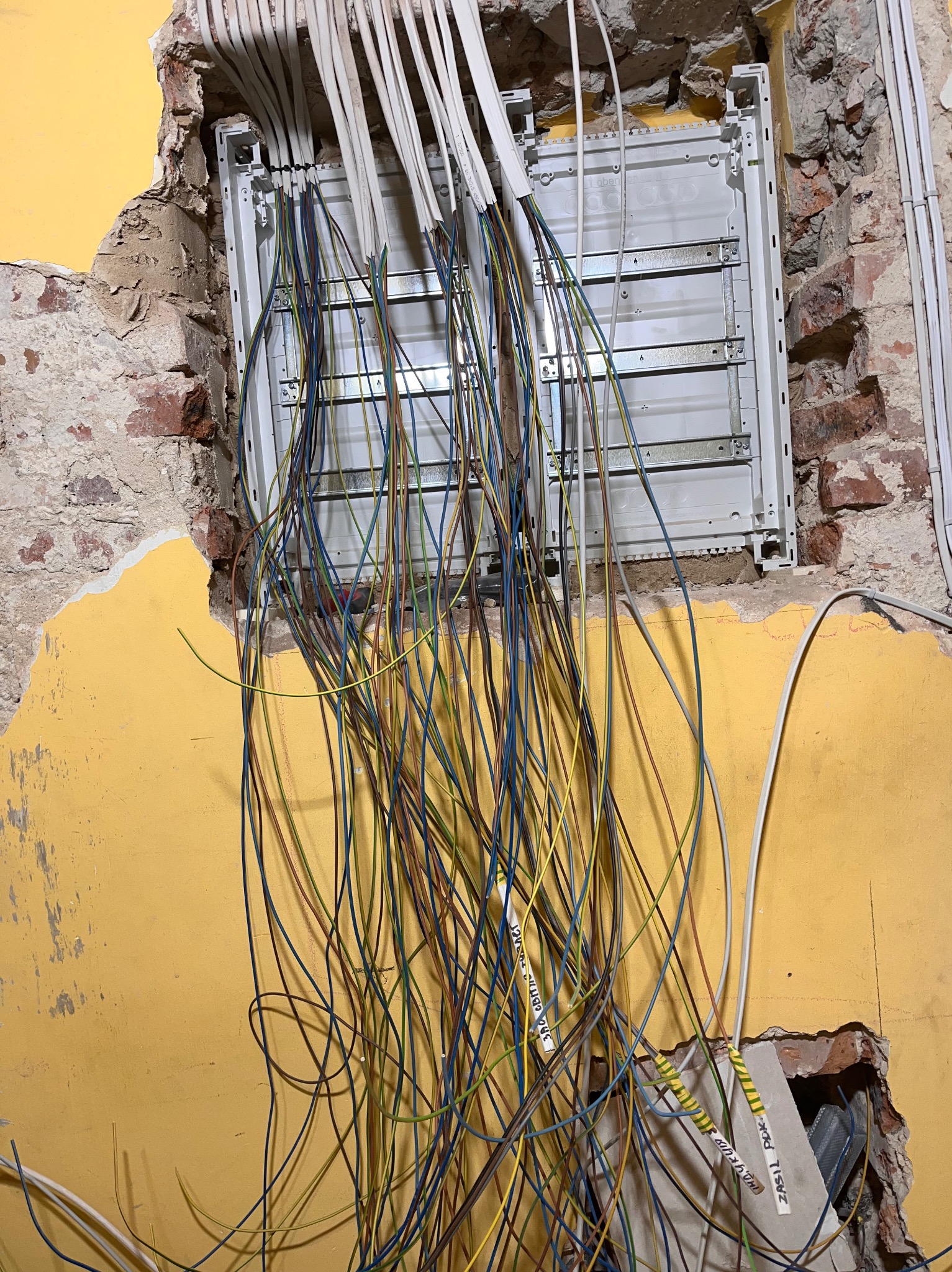 Rozdzielnia elektryczna w trakcie montażu w ścianie z żółtą farbą i odsłoniętą cegłą, z widocznymi przewodami w kolorach niebieskim, zielonym i brązowym, oraz białymi kablami w listwach.