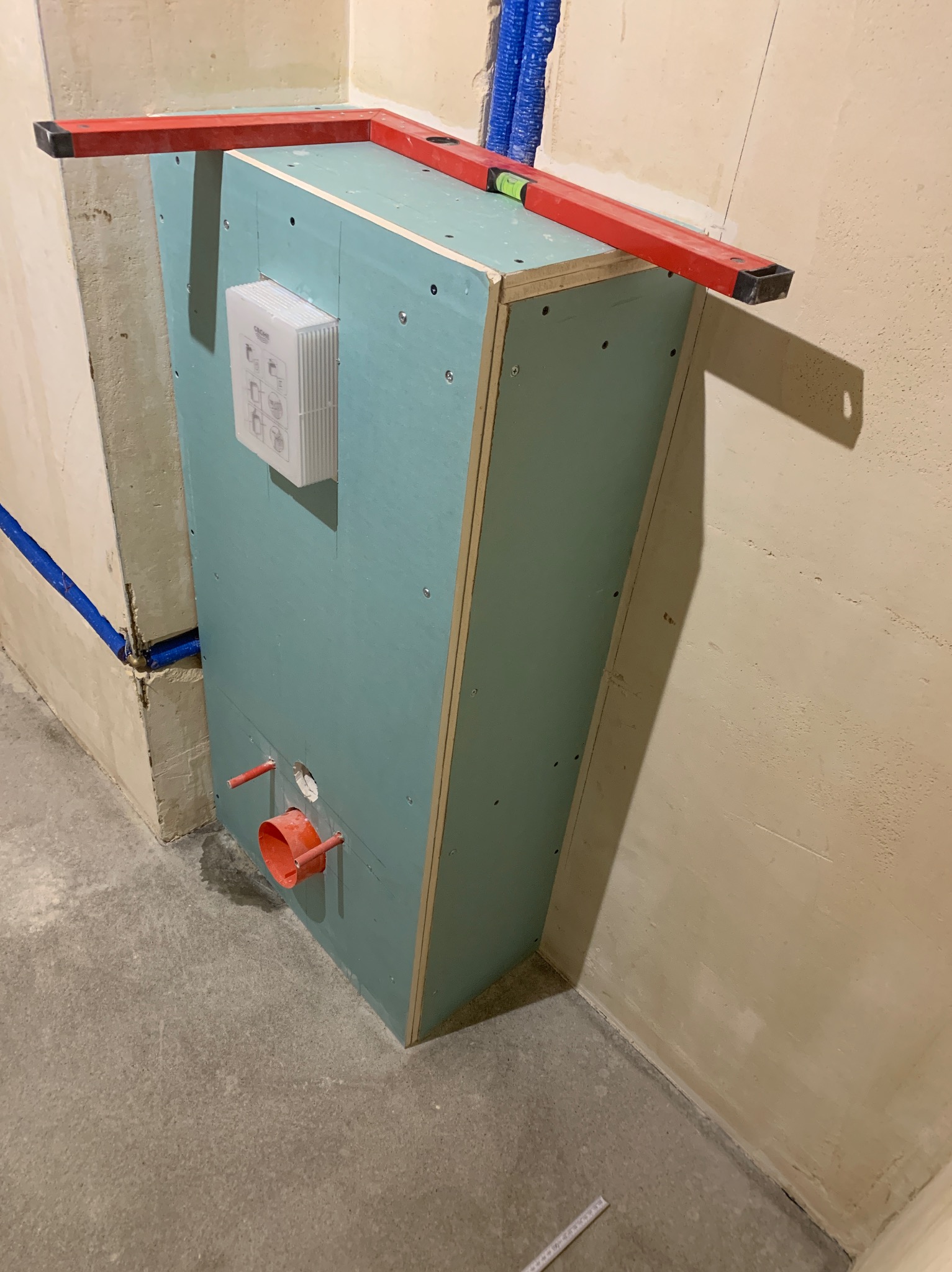 Stelaż podtynkowy zabudowany płytą gipsowo-kartonową, poziomowany czerwoną poziomicą, widoczne przyłącza kanalizacyjne i wodne, biały panel sterujący z instrukcjami