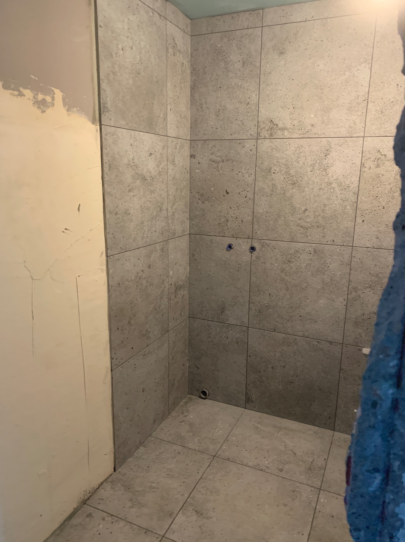 Wnętrze łazienki w trakcie remontu, widoczne szare, kwadratowe płytki na ścianach i podłodze, surowa ściana z widocznymi śladami po gładzi, dwa otwory na baterię w ścianie, odpływ w podłodze.